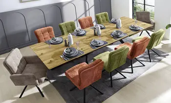 Langer Esstisch aus Eiche mit acht gepolsterten Stühlen in Beige, Orange und Grün, eingedeckt mit grauen Tellern, Gläsern und Stoffservietten.